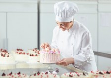 A female confectioner is holding a cake in her hand and in another hand is showing the ok sign in the bakery.